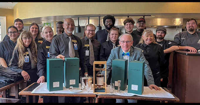A group of people standing together in 1875: The Steakhouse at French Lick Springs Hotel, posing behind a table displaying bottles of Release No. 37: The Last Drop 27 Year Old Kentucky Straight Bourbon from Buffalo Trace Distillery. Some in the group are wearing uniforms or name tags, and one person is seated, smiling, wearing a colorful bow tie. The background features kitchen equipment.