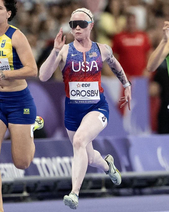 Athlete in USA uniform running on track, wearing sunglasses, with competitors in background at Paris 2024 event.