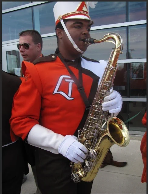 A person in a marching band uniform is playing a saxophone. The uniform is red, black, and white with a distinctive emblem on the chest. They are standing outside near a glass building. Another individual in sunglasses is visible in the background.