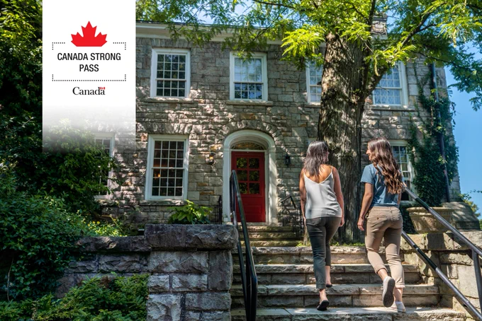 Two women walking up stone steps toward a historic building with a red door surrounded by greenery on a sunny day.

Top left corner: Canada Strong Pass logo and Canada wordmark