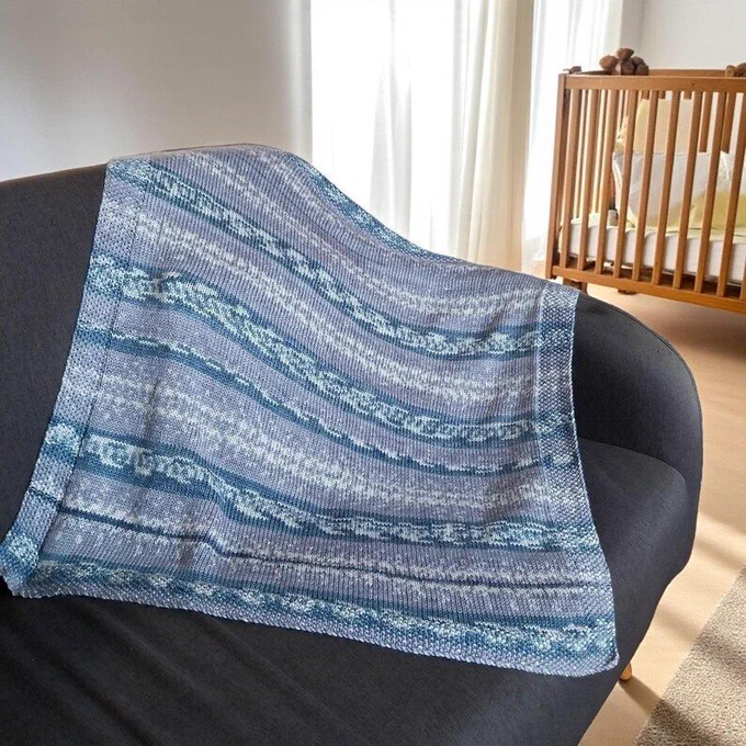 A hand knitted grey and white striped baby blanket draped over a dark sofa, with a wooden crib and white curtains in the background.