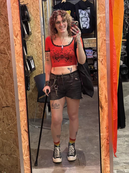 A picture of me taking a mirror selfie in a shop, wearing black shorts, a red cropped tshirt, black converse and rainbow socks. I am also holding a cane and a bag.
