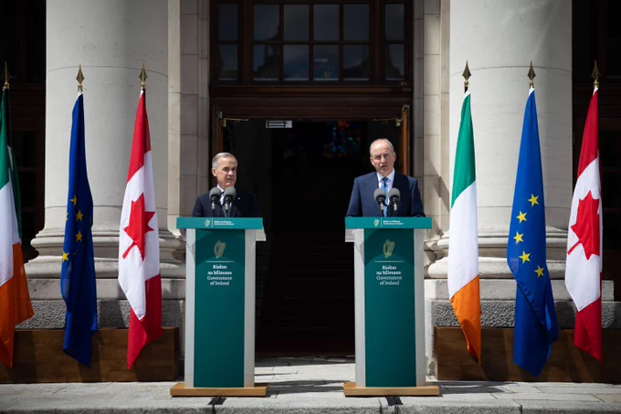 Prime Minister Mark Carney and the Taoiseach of Ireland, Micheál Martin, are standing behind podiums and delivering a joint media availability in Dublin, Ireland. The flags of Ireland, the European Union, and Canada are situated next to them.