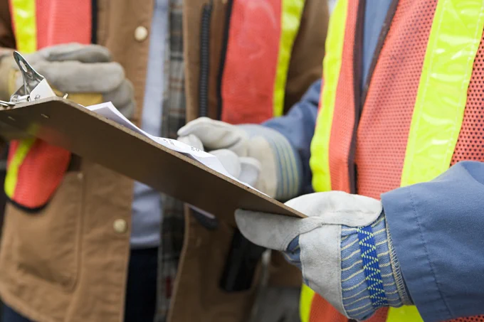 Construction workers wearing safety vests reviewing documents on a clipboard at a job site.