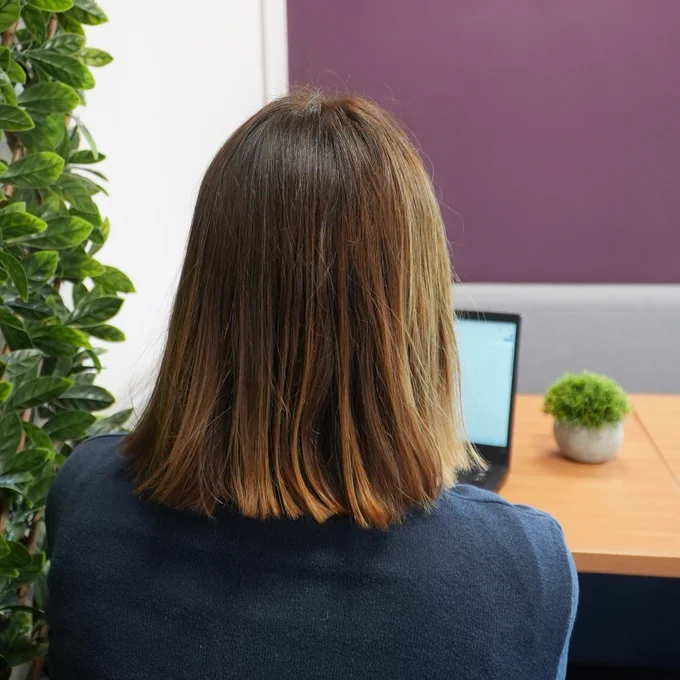 Woman with shoulder-length brown hair working on a laptop at a desk, with plants in the foreground and background.