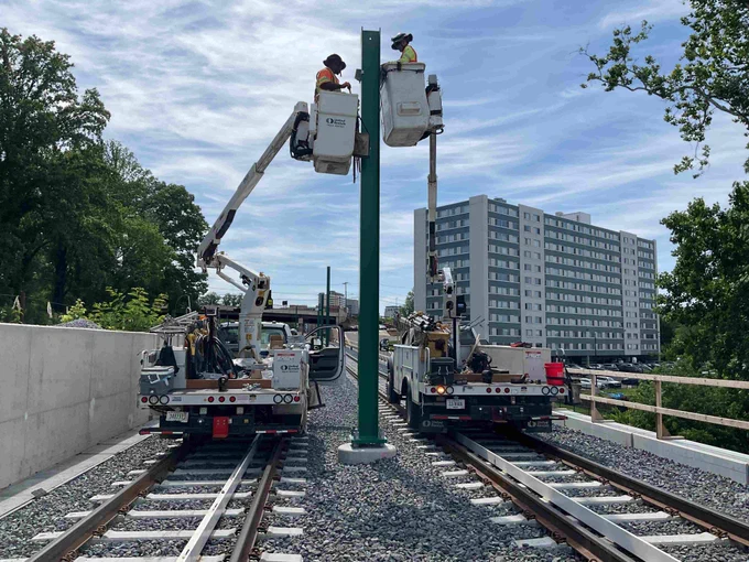 Two workers in bucket trucks installing or maintaining a tall green pole between parallel train tracks under a partly cloudy sky.