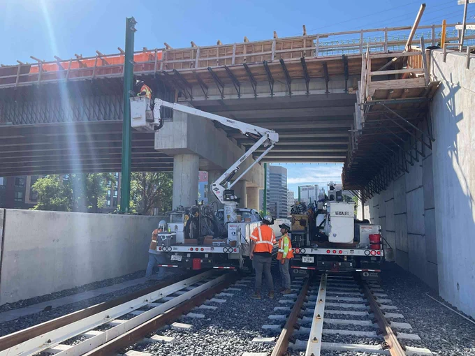 Workers in safety gear operate machinery on railway tracks beneath a bridge under construction on a sunny day.
