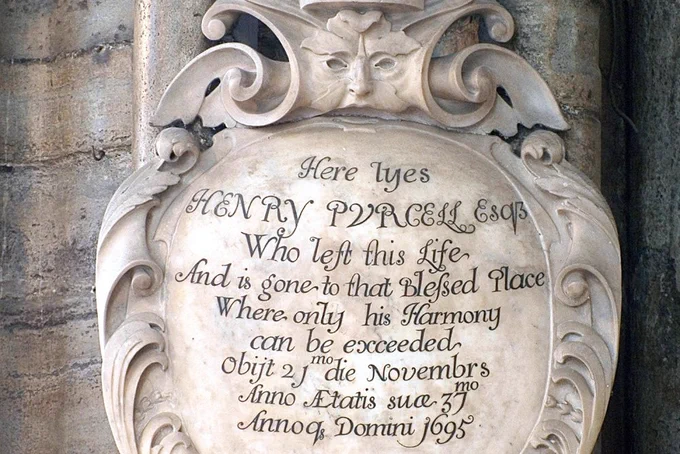 An ornate, carved white marble plaque commemorating Henry Purcell, decorated with elegant scrollwork and a mask-like face at the top. The inscription reads: 'Here lyes Henry Purcell Esq., who left this life and is gone to that blessed place where only his harmony can be exceeded. Obijt 21st die Novembris Anno Aetatis suae 37 Anno Domini 1695.'