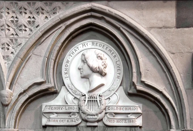 A stone relief set in an ornate arched frame, featuring a profile bust of Jenny Lind with braided hair. Surrounding the bust is an inscription: "I know that my Redeemer liveth." Below, a lyre is carved, along with the name "Jenny Lind Goldschmidt" and the dates "Born Oct 6, 1820" and "Died Nov 2, 1887." The background displays intricate floral patterns.