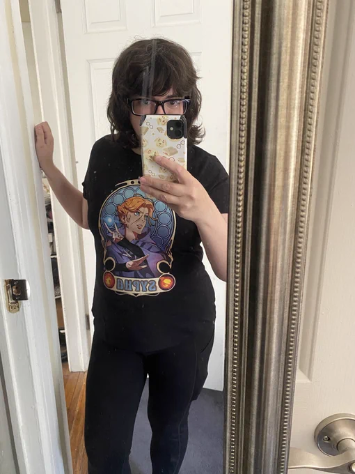 A photo of me, a white woman with brown hair taking a selfie in the mirror, I’m wearing a Sypha Belnades Castlevania shirt