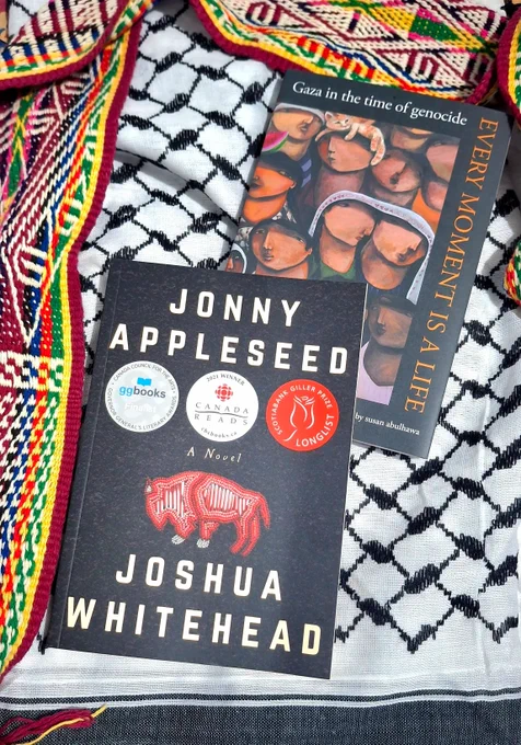 A photo of two books. One is Jonny Appleseed by Joshua Whitehead, with the cover black and adorned by an artwork of a red bison studded with glitter. The other is Every Moment is a Life: Gaza in the Time of Genocide. The cover depicts a painting of Palestinian refugees, one has an orange cat on their head. The books are presented on a keffiyeh and a colorful woven scarf.