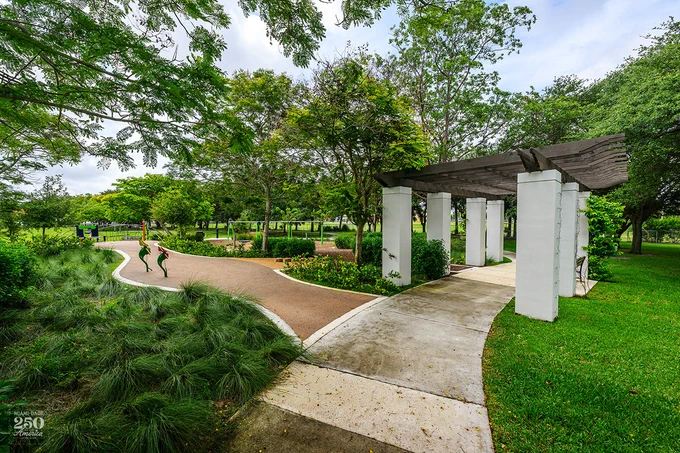 A lush park scene features a winding brown path with abstract green & red sculptures, leading to a playground in the distance. On the right, a concrete path passes under a white-columned pergola. Green trees & grass fill the landscape. Text: MIAMI-DADE 250 America.