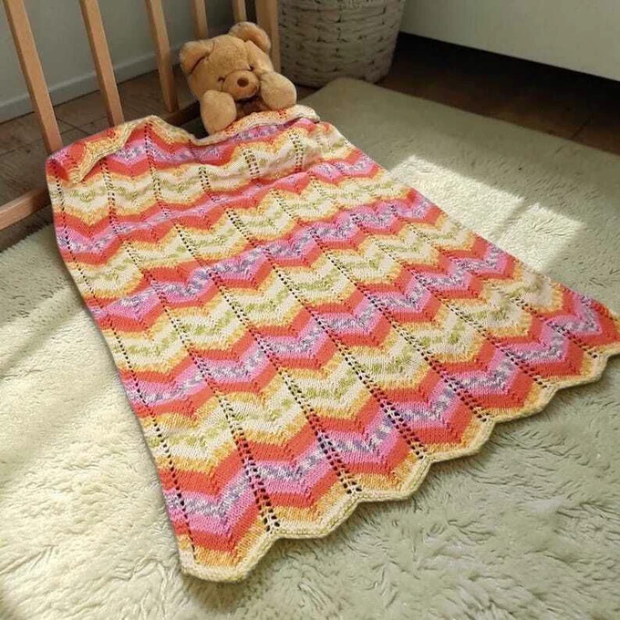 Knitted blanket spread on fluffy cream rug, featuring vibrant chevron pattern in orange, pink, yellow and green with scalloped edge. Brown teddy bear peeks from top corner near wooden crib rails. Woven basket and plant in background. Warm natural light highlights delicate lace texture and bright colours.