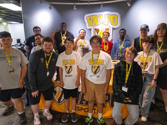 A group of Valpo Esports players and coaching staff smile beside younger students inside Valparaiso University's Esports Lab during Valpo Esports Summer Camp.