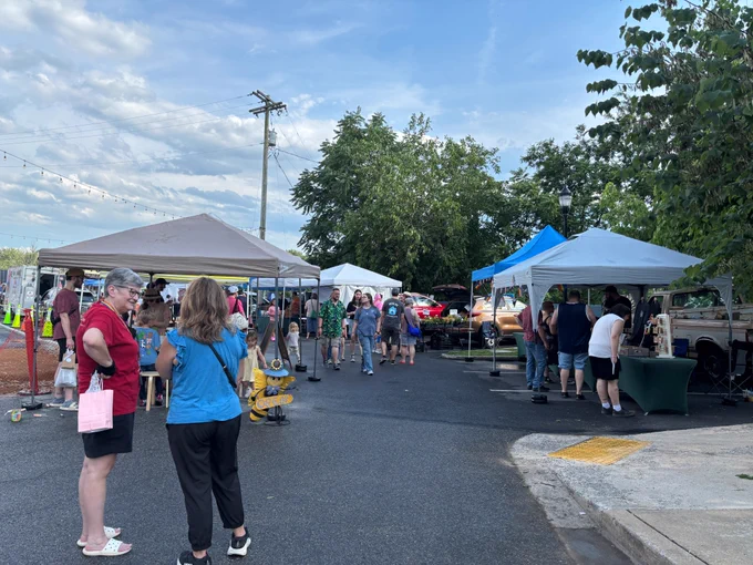 An outdoor street market scene with several pop‑up tents arranged along a paved roadway. The tents are in different colors, including tan, white, and blue. Under the tents, vendors display various items, and groups of people browse the booths. In the foreground, two individuals stand near a tan tent; one is holding a pink shopping bag while another stands with a backpack. A yellow fire hydrant decorated with nautical‑themed props is positioned near the walkway. More people are visible walking through the market toward the center of the image. In the background, trees line the area, and cars—including a red vehicle—are parked behind the tents. Overhead, string lights are faintly visible against a partly cloudy sky.