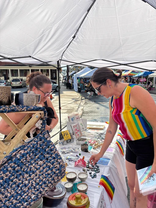 A scene at an outdoor market under a white canopy tent. A display table is covered with a white cloth and arranged neatly with handmade items, including candles in decorative containers, small painted stones, framed art prints, pamphlets, and other craft goods. Several small rainbow flags are placed along the table’s edge. In the foreground, an individual wearing a brightly colored rainbow‑striped sleeveless top and dark shorts is reaching toward items on the table while holding a stack of brochures or cards. Another individual stands behind the table, partially visible, surrounded by hanging woven bags and additional merchandise. In the background, beyond the tent, a street lined with buildings and more vendor tents can be seen, with people browsing and walking through the market.