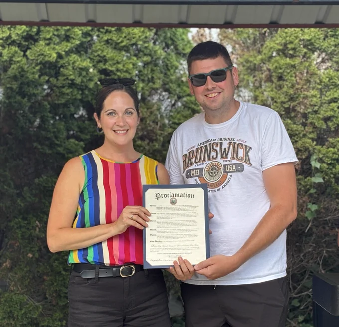 Two individuals stand outdoors in front of dense green shrubbery under a metal-roofed structure. They are holding a framed proclamation document together at the center of the image. The individual on the left is wearing a sleeveless, vertically striped top in bright colors including red, blue, yellow, and green, paired with dark pants and a belt. The individual on the right is wearing a light-colored T‑shirt with a printed design that includes the word “BRUNSWICK” and other text elements. Both individuals hold the proclamation at its edges, with the document featuring a bold title at the top and several paragraphs of printed text beneath it. The background includes leafy plants and shadows cast from the overhead roof. 