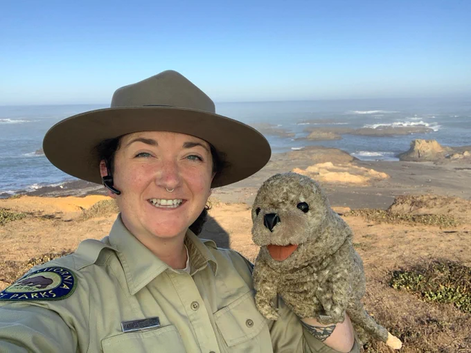 California State Parks Interpreter posing in front of the ocean coast in uniform with sea lion stuffed animal.