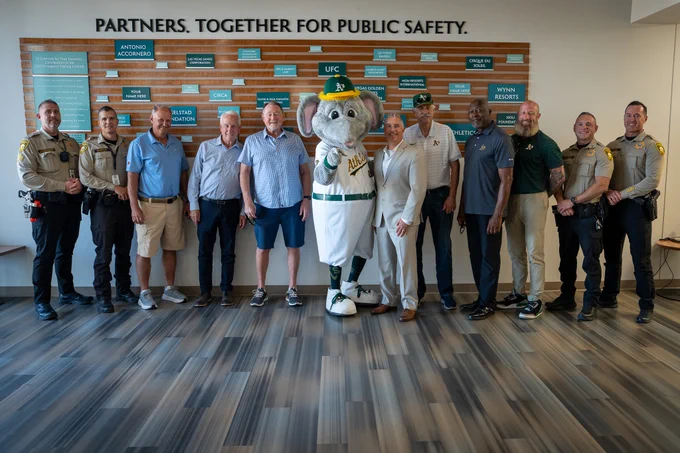 Group of law enforcement officers and officials standing with a mascot in front of a public safety partnership wall.