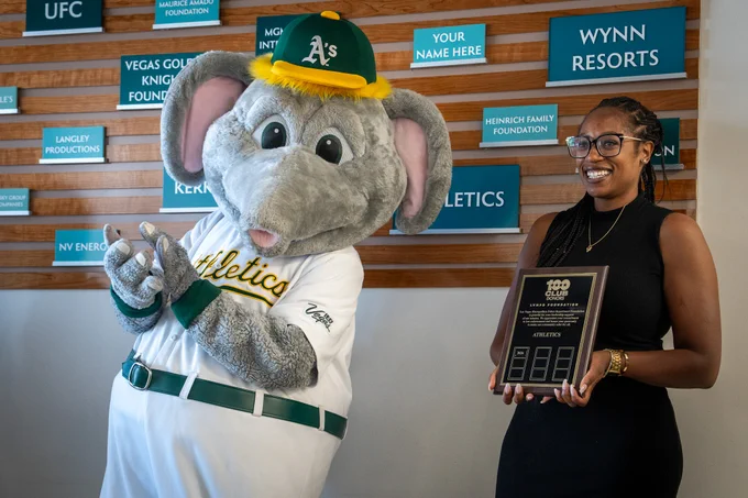 Oakland Athletics mascot Stomper poses with a person holding a black award plaque in front of a wooden wall with sponsor signs.