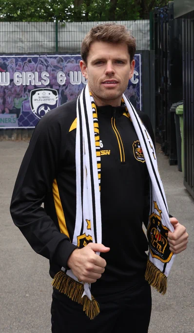 A man stands outdoors wearing a black and yellow sports jacket and holding a football scarf. He looks confidently at the camera, conveying a proud and energetic mood.