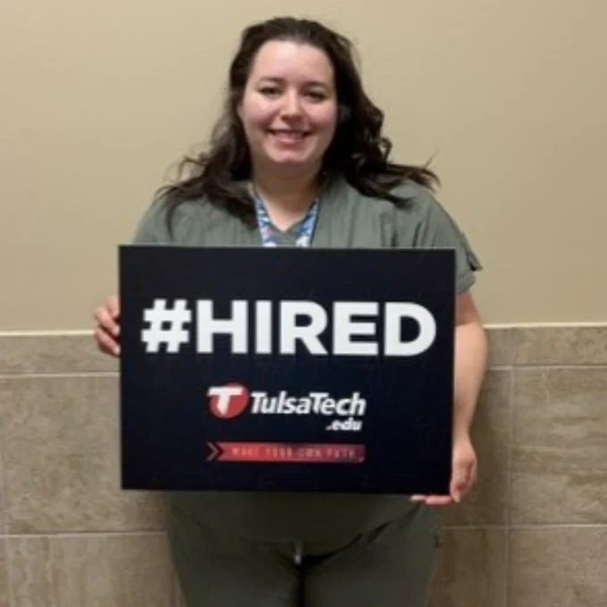 Person holding a #HIRED sign from Tulsa Tech against a beige tiled wall.