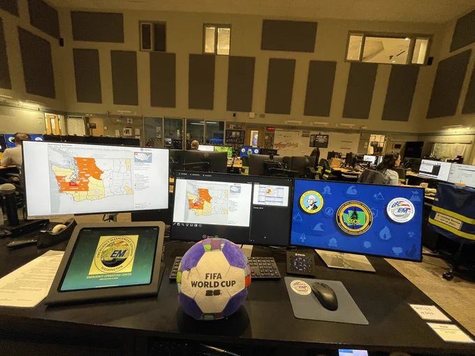 A large room with multiple computers and monitors is seen. The backs of people’s heads as they work. A map of Washington state with some counties highlighted on one computer. Logos on another. A plush soccer ball with FIFA World Cup sits on a table.