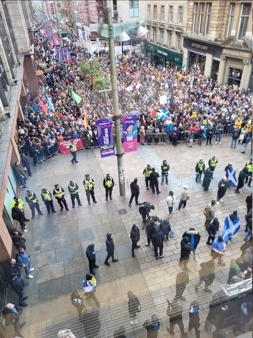 A high-angle view of a crowded street protest in a city center. Thousands of people stand behind metal barriers holding flags and signs, while a line of police officers in yellow jackets forms a cordon in the foreground. Scottish and Palestinian flags are visible among the crowd.