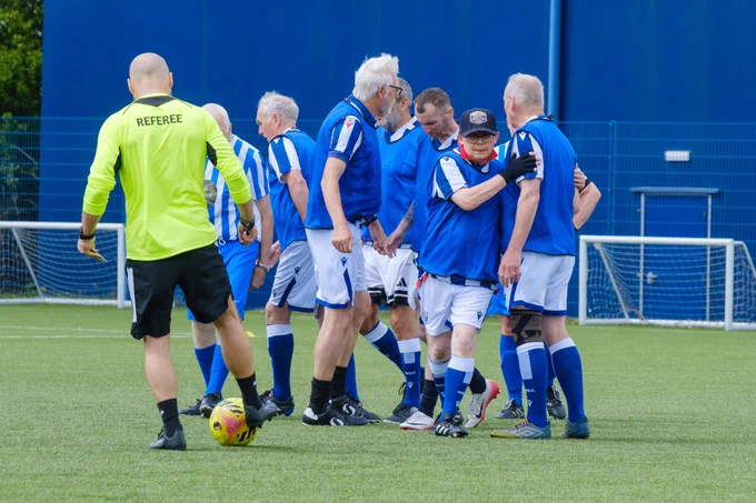 Group of Cancer Kickers players in blue-and-white kits cluster together celebrating a goal, while a referee in a yellow shirt walks past a stationary ball in the foreground.