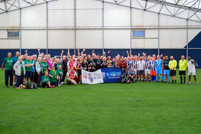 Tournament participants and spectators gathered on the pitch for a photo. Moore than Cancer and Albion Foundation banners are held at by people at the front.