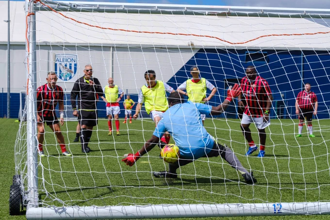 Action shot from behind the goal net as a goalkeeper in a light blue top dives low to stop a close-range shot, while several players in red-and-black and yellow bibs watch the ball near the goal line on a sunny pitch.