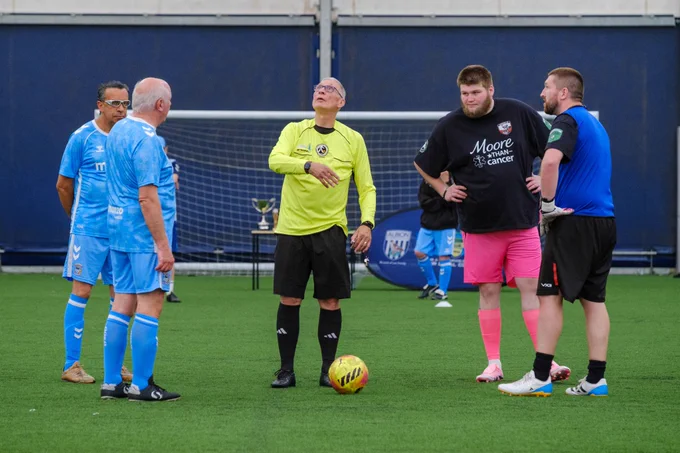 Referee in a bright yellow shirt stands on an outdoor pitch with a football at their feet, speaking to two small groups of players in blue and dark kits gathered for a pre-match coin toss, with a goal and trophy table visible behind.