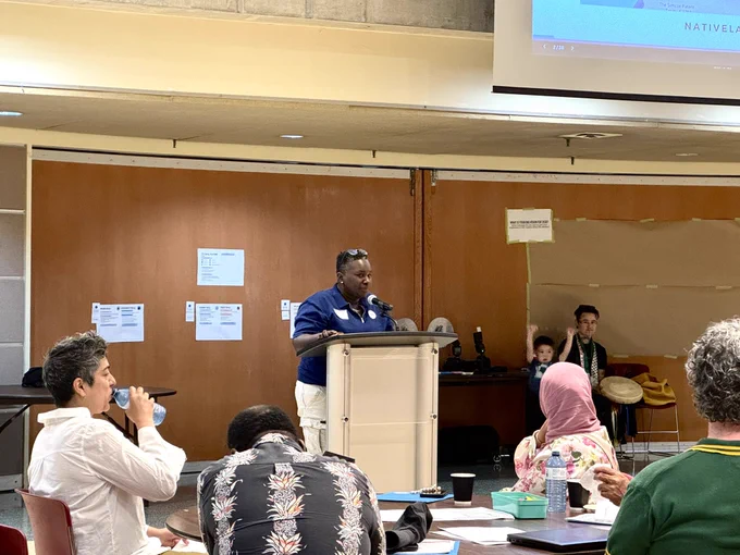 Stacia opens the summit with a Land acknowledgement. She stands at the podium and is seen facing the room and 4-5 participants are seen watching her.