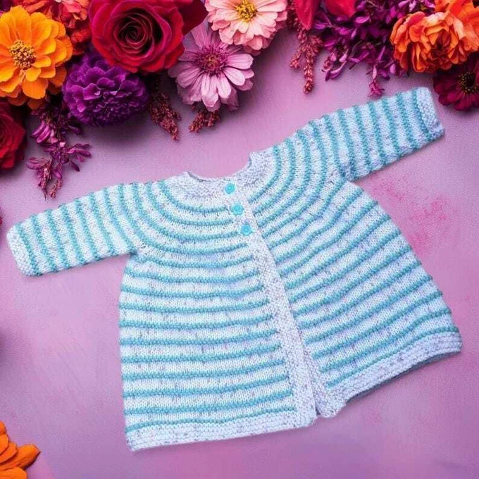 Knitted baby cardigan laid flat on soft pink background. Features fine horizontal stripes in pale green and white, with round neck and small blue buttons. Top edge framed by vibrant bouquet of red roses, orange marigolds, pink dahlias and purple blooms. Bright light highlights delicate knit and vivid colours.