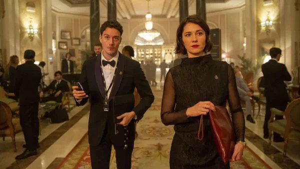 A still from the film Rich Flu. An elegantly dressed, irritated-looking white woman carrying a slender leather briefcase walks through an expensive hotel lounge followed by a smartly dressed but subservient-lookimg white man.