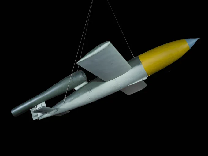 V-1 flying bomb, also known as a buzz bomb, suspended against a black background. It is light green with a bright yellow band around the front of it.
