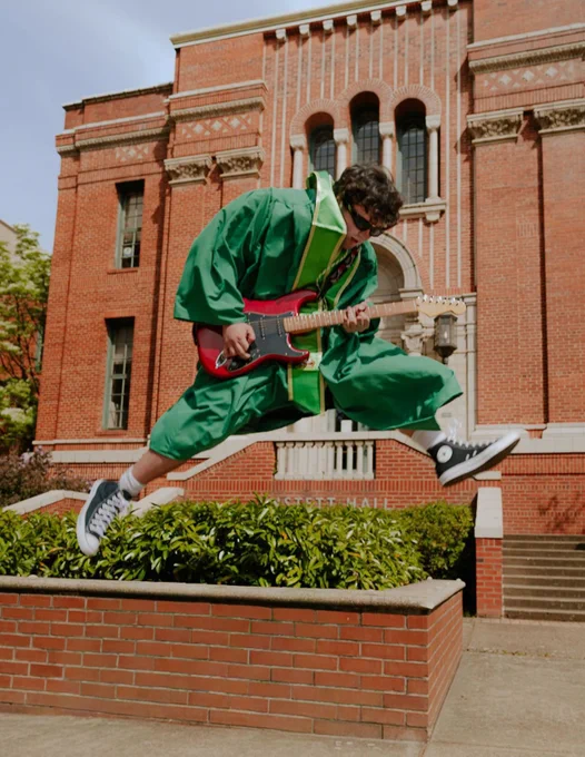 A person in a green graduation gown jumps while playing a red electric guitar in front of a brick building. 