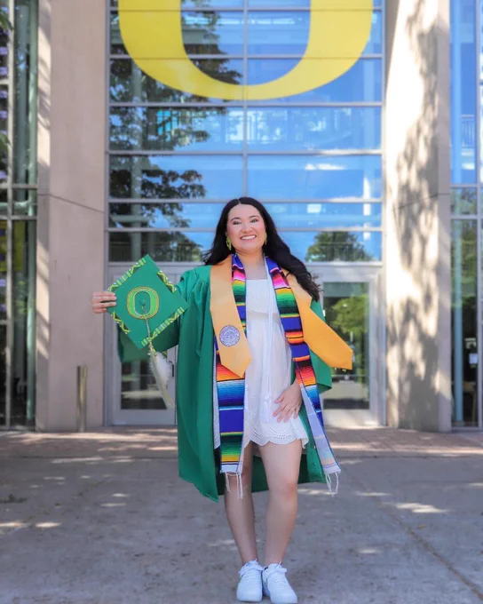 A person in graduation attire holds a decorated cap with the University of Oregon's "O" logo. They stand in front of a building with a large "O" symbol in the background.