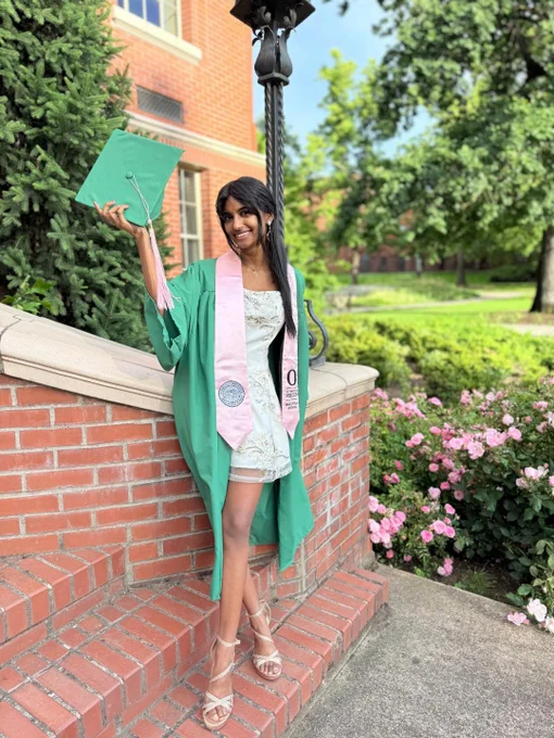 A person in a green graduation gown and cap stands on brick steps, holding up the cap with one hand. They are wearing a pink stole and smiling. A lush garden with pink flowers and trees is in the background.