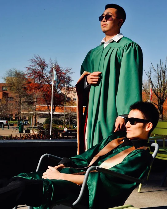 Two individuals in green graduation gowns are outdoors. One is standing with hands clasped, and the other is seated, both wearing sunglasses. The setting includes a backdrop with trees and buildings.