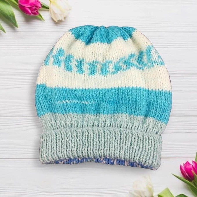 Knitted beanie on white wood, with horizontal bands in cream, bright turquoise and pale mint green. Features a decorative turquoise pattern across the cream section. Folded ribbed brim. Tulips frame corners. Bright light highlights texture and colour contrast.