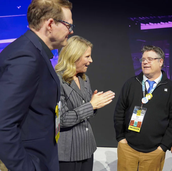 Three conference attendees engaged in conversation, wearing badges and standing in a dimly lit event space.