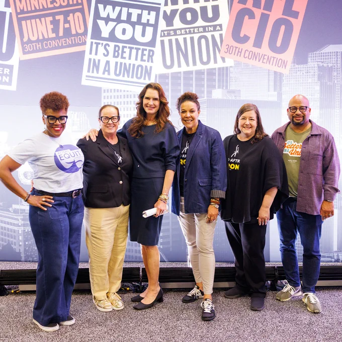 Group of six diverse women standing together at a union convention with signs promoting unity and equity in the background.