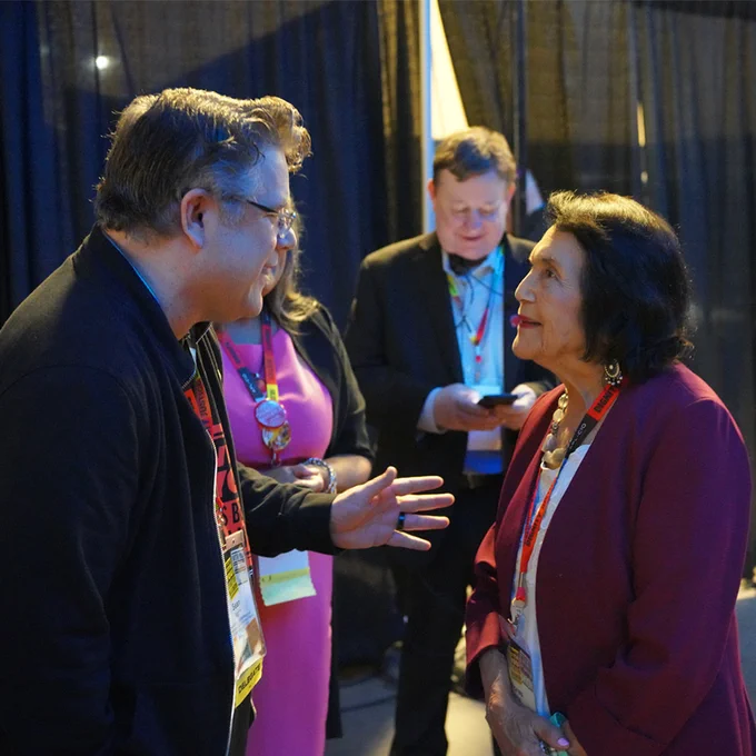 Two people engaged in a focused conversation at an indoor event with black curtains in the background.
