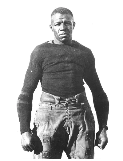 Black and white photo of Duke Slater wearing a tight, long-sleeved shirt and pants, standing with clenched fists against a plain white background.