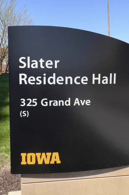 Black sign with white text reading 'Slater Residence Hall, 325 Grand Ave (S)' and the word 'IOWA' in yellow at the bottom, set outdoors with grass and trees visible.