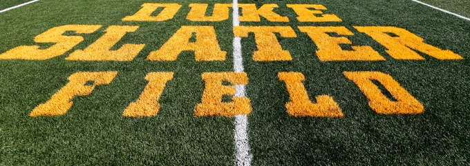 Green artificial turf with large yellow letters spelling 'DUKE SLATER FIELD' centered on the field