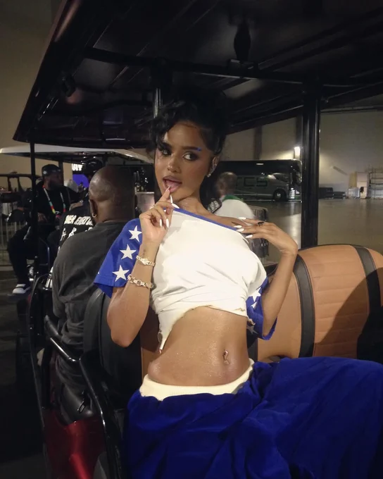A person sits confidently in a vehicle, wearing a cropped American flag-themed outfit and holding a lollipop. The setting has a casual and playful atmosphere.
