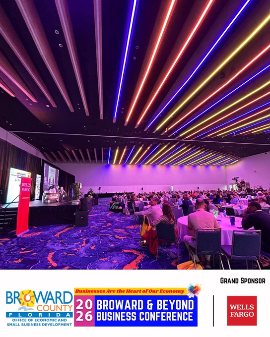 A large conference room at the Broward County Convention Center filled with attendees seated at tables. The room features colorful, illuminated ceiling lights and multiple display screens. Banners for the2026 Broward & Beyond Business Conference and Grand Sponsor Wells Fargo are visible.