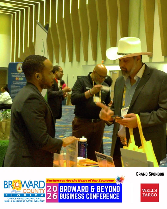 Two people are conversing at a business conference. One is wearing a suit and holding a bag, with a white cowboy hat. The other is dressed in a dark jacket and appears engaged in discussion. Background attendees are interacting near a booth. The event is the 2026 Broward & Beyond Business Conference, Grand sponsor is Wells Fargo.